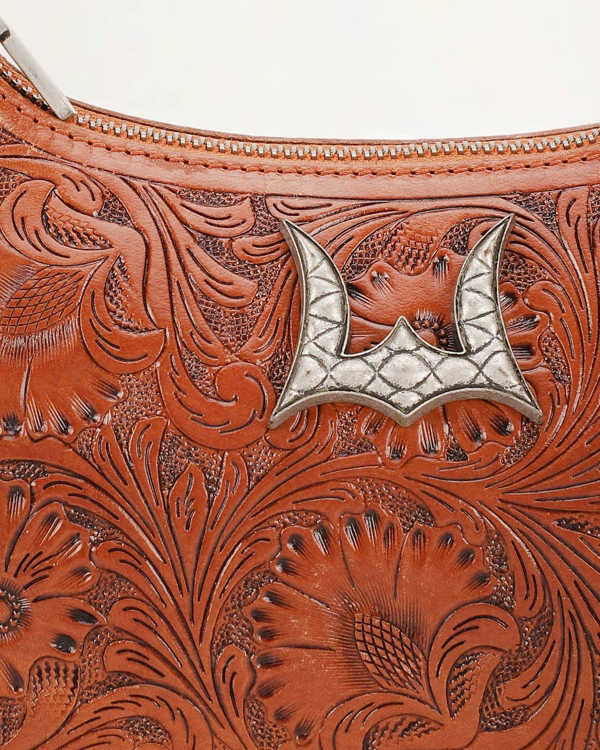 Close-up of a brown tooled leather handbag with floral embossing and a silver bat-shaped metal emblem near the zipper.