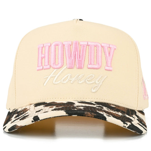 Front view of full-cotton trucker cap featuring pink embroidered “Howdy” and off-white embroidered “Honey” lettering across the cream crown, a black and white cow print curved brim, and playful western-inspired country styling.
