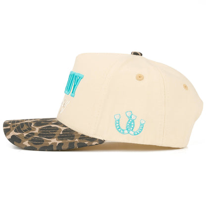 Side view of cream full-fabric trucker cap with turquoise horseshoe embroidery on the side panel, “Howdy Honey” embroidery visible from the front, black and white cow print brim, and fun feminine western detailing.