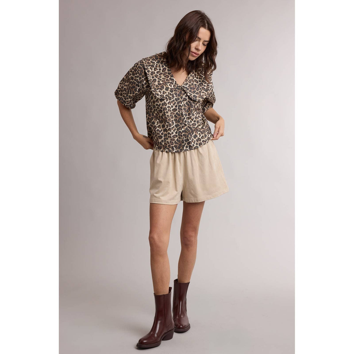 Woman wearing a leopard print top and beige shorts with brown boots on a plain background