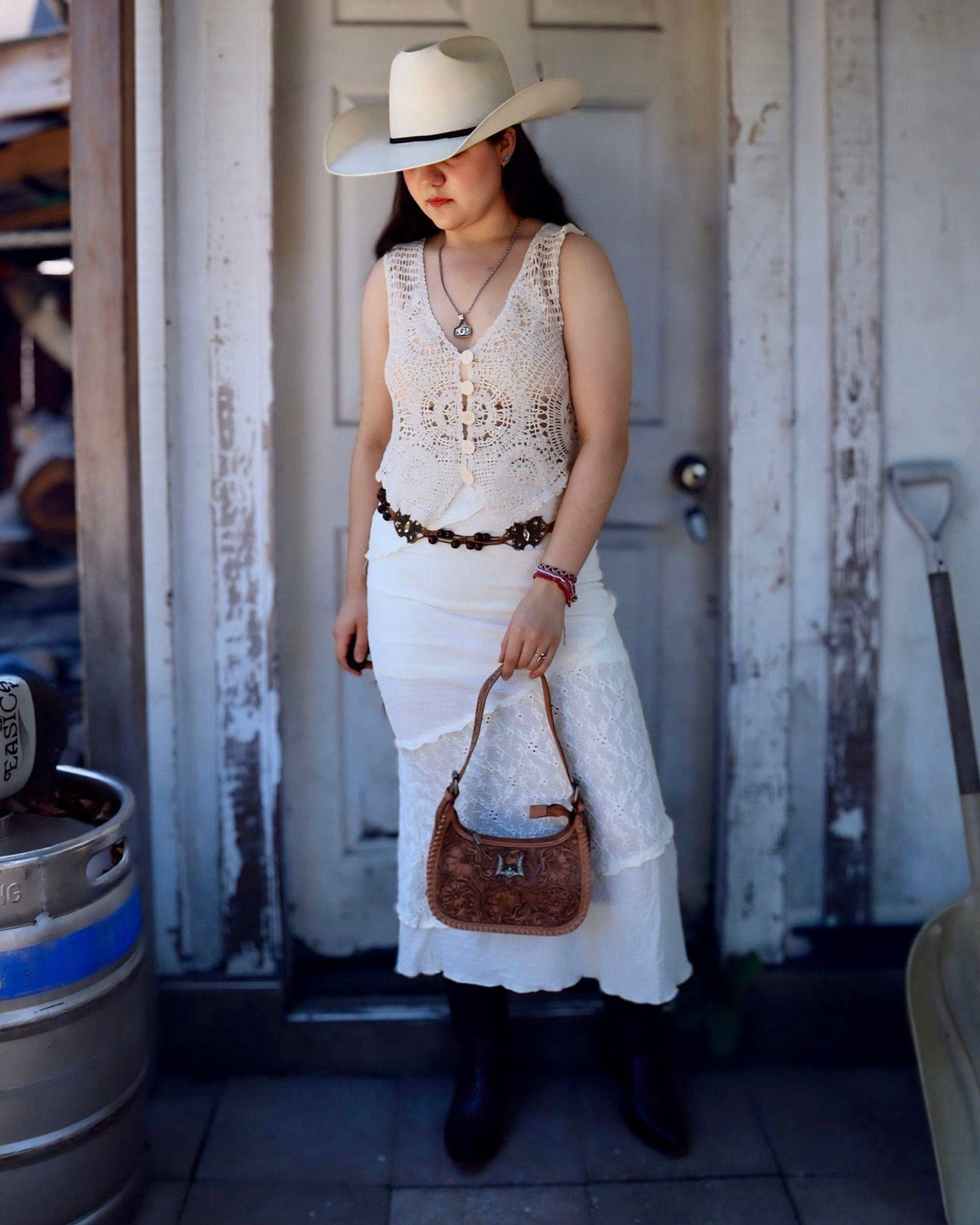 A woman stands in front of a weathered white door wearing a cream crochet vest, a white skirt, a brown beaded belt, a white cowboy hat, and black boots, while holding a brown leather handbag.